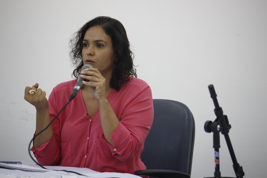 Na foto, Ana Paula está sentada e fala segurando o microfone, enquanto a outra mão está com o punho fechado. Ela usa uma blusa rosa de botão.